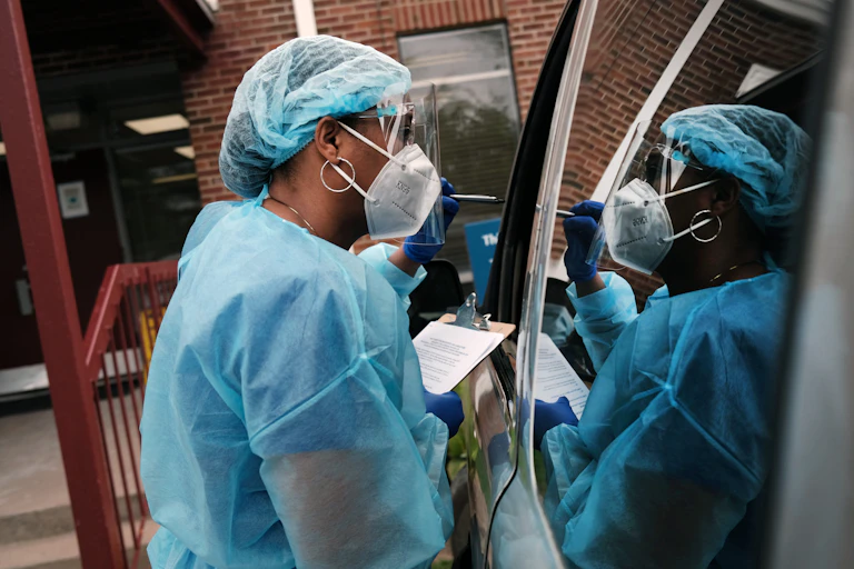 A worker wearing a mask and other personal protective equipment and carrying a clipboard speaks with passengers in a parked vehicle.
