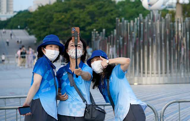 Three Tokyo Olympics volunteers posing for a selfie in front of the Olympic flame
