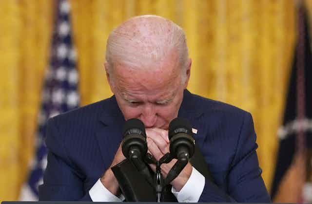 Joe Biden stands a lectern in the White House with his eyes closed and his hands clasped.