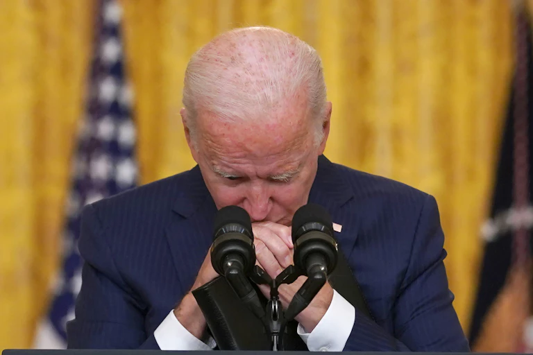 Joe Biden stands a lectern in the White House with his eyes closed and his hands clasped.