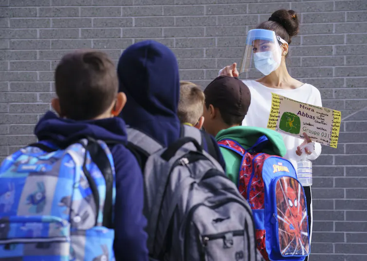 A teacher in face shield faces students in a yard.