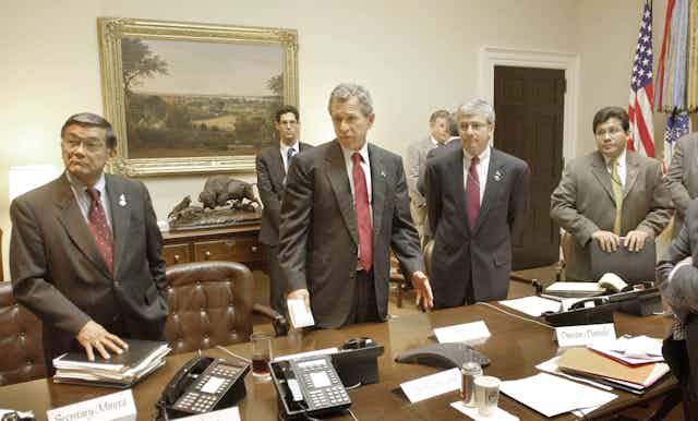 Men in suits stand around a meeting table
