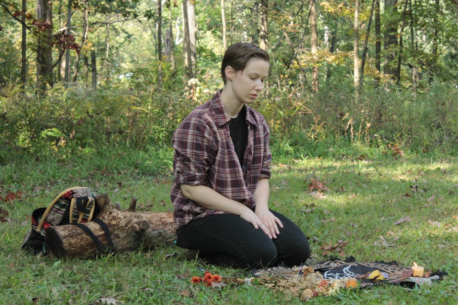 A person in a flannel shirt and black pants kneels in the grass in front of a Wiccan altar.