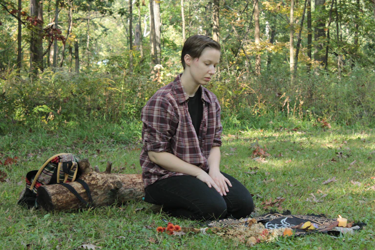A person in a flannel shirt and black pants kneels in the grass in front of a Wiccan altar.