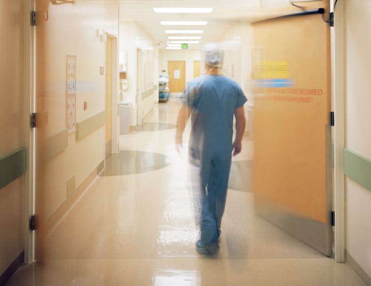 Surgeon in hat and blue scrubs walking in hospital corridor away from camera