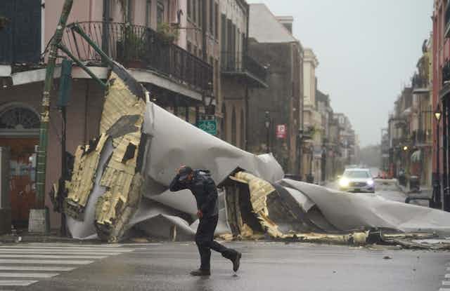 A man leans against the wind, holding his baseball cap on his head as he passes the mangled wreckage of a roof now in the street.