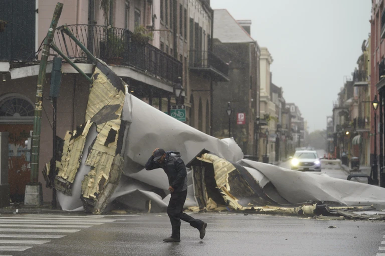 A man leans against the wind, holding his baseball cap on his head as he passes the mangled wreckage of a roof now in the street.