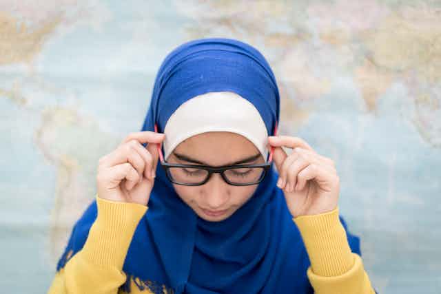 A young female Muslim student wearing a blue headscarf adjusts her glasses while standing in front of a world map.
