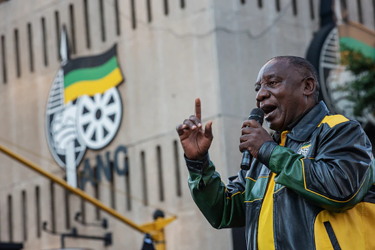 A man speaks into a microphone held in his left hand, while his right index finger points skywards.