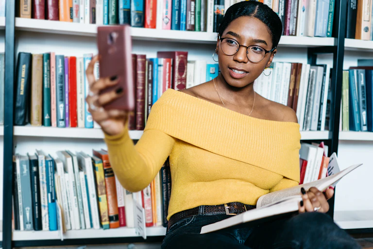 A woman in a yellow top and glasses holds a mobile phone with a book on her lap