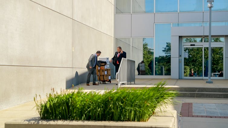 Men in suits move large boxes of documents around outside a courthouse.