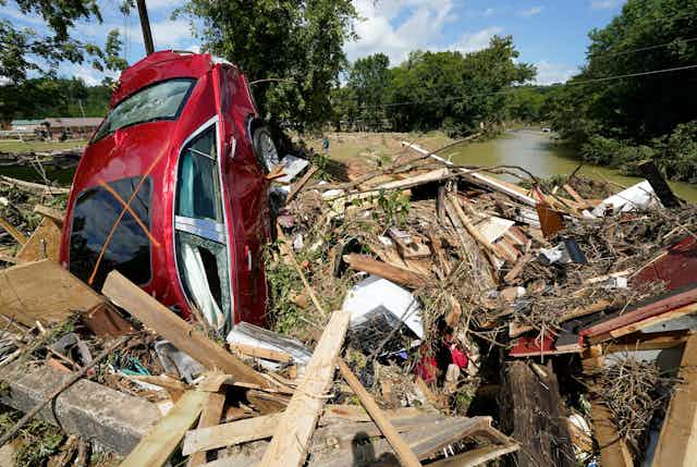 A car with broken windows is nose down in debris, half buried and partial in a river.