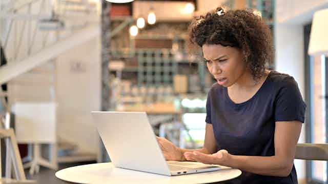 A young woman in a black shirt looking at her laptop with an expression of disbelief.