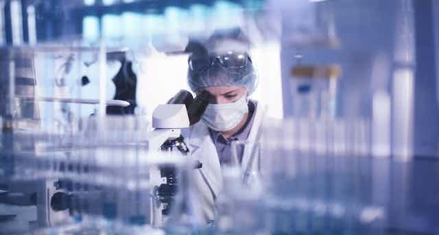 A woman wearing a hair mask and a mask looks into a microscope with vials in the foreground