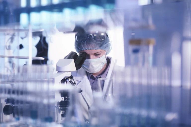 A woman wearing a hair mask and a mask looks into a microscope with vials in the foreground
