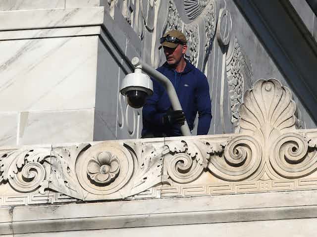 A man standing on a ledge on the exterior of a marble building holds a pole with a security camera at one end