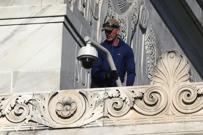A man standing on a ledge on the exterior of a marble building holds a pole with a security camera at one end