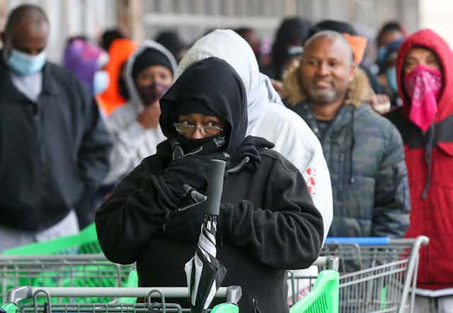 Shoppers clearly cold in their light jackets wait to get into a store. The woman in front has hood and scarf covering most of her face.
