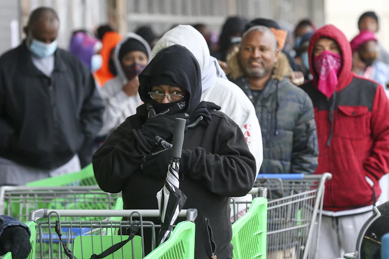 Shoppers clearly cold in their light jackets wait to get into a store. The woman in front has hood and scarf covering most of her face.