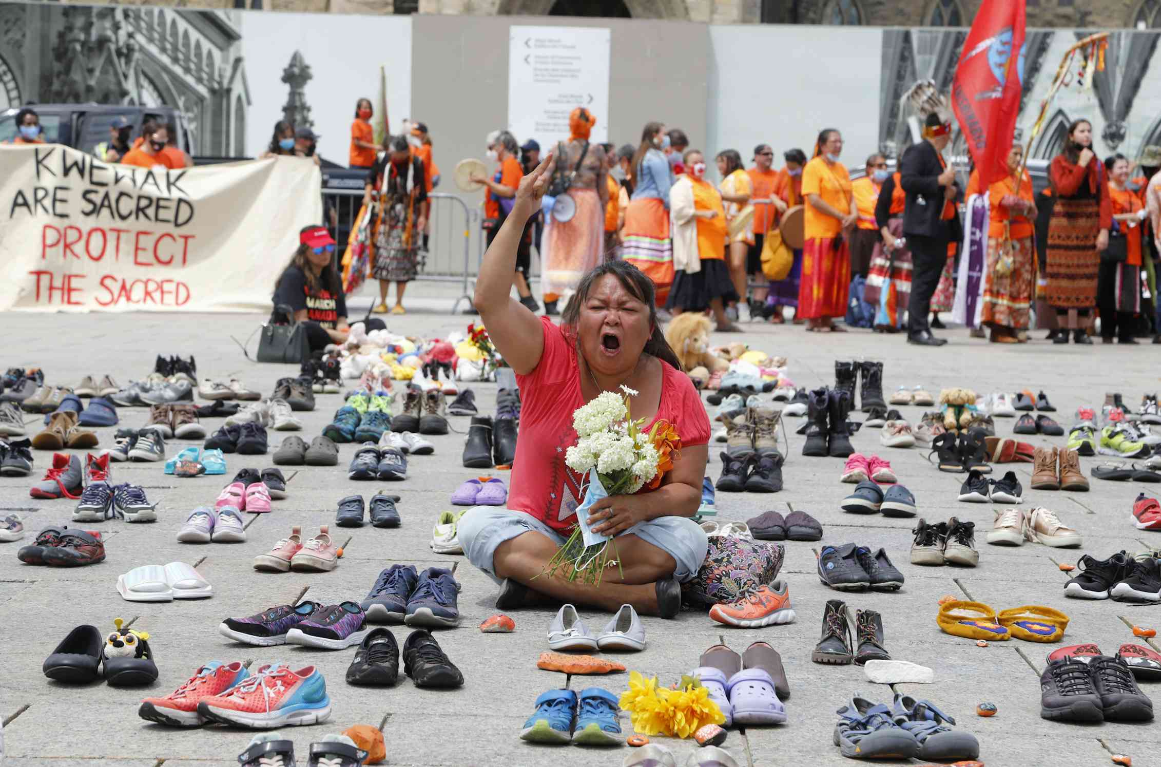 Woman sits with fist in the air, wearing orange t-shirt, holding teddy bear, surrounded by hundreds of little shoes