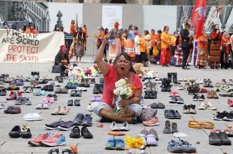 Woman sits with fist in the air, wearing orange t-shirt, holding teddy bear, surrounded by hundreds of little shoes