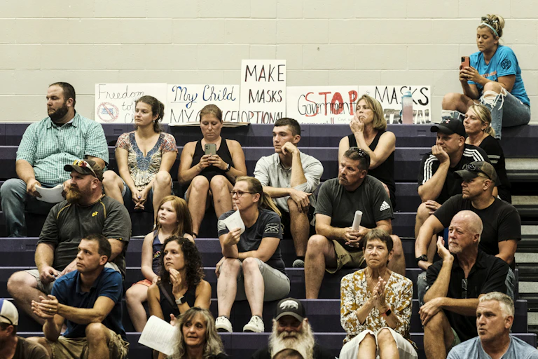 A crowd of parents sit on bleachers next to anti-mask signs.