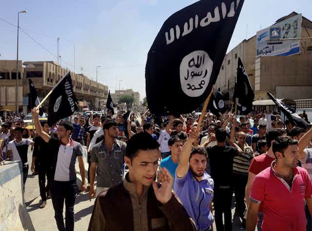 A crowd of people fill a street waving black Islamic State group flags.