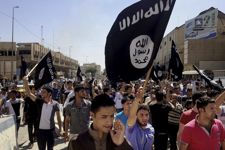 A crowd of people fill a street waving black Islamic State group flags.