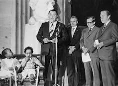 A man speaks on stage next to two Black children and a group of other men.