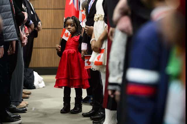 Une fillette vêtue d'une robe rouge, entourée d'adultes, tient un drapeau canadien