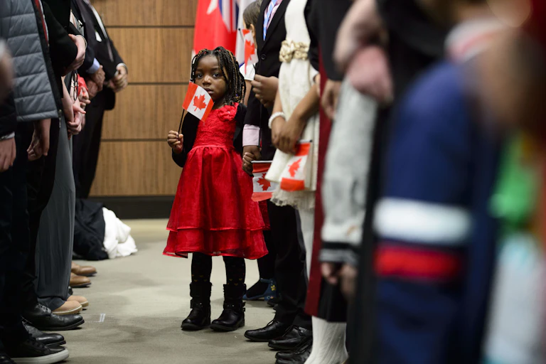 Une fillette vêtue d'une robe rouge, entourée d'adultes, tient un drapeau canadien