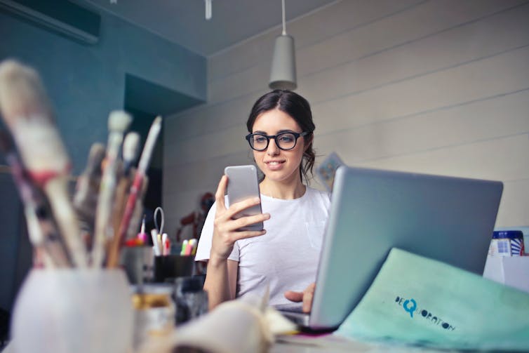 Woman sitting at laptop, wearing glasses looks at cellphone