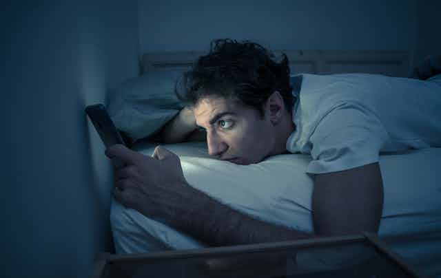 Man lays in bed with the lights off, the glow of his cellphone illuminates his face