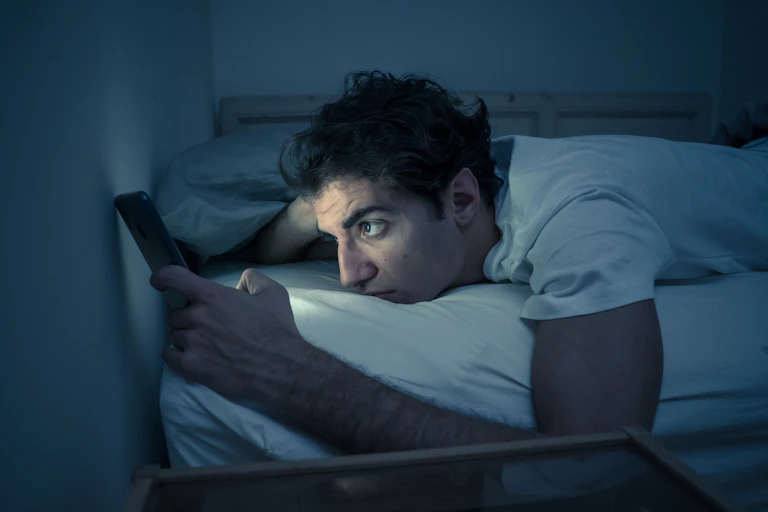 Man lays in bed with the lights off, the glow of his cellphone illuminates his face