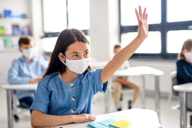 Girl sitting at school desk wearing a mask, with her hand up.