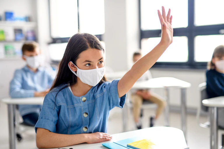Girl sitting at school desk wearing a mask, with her hand up.