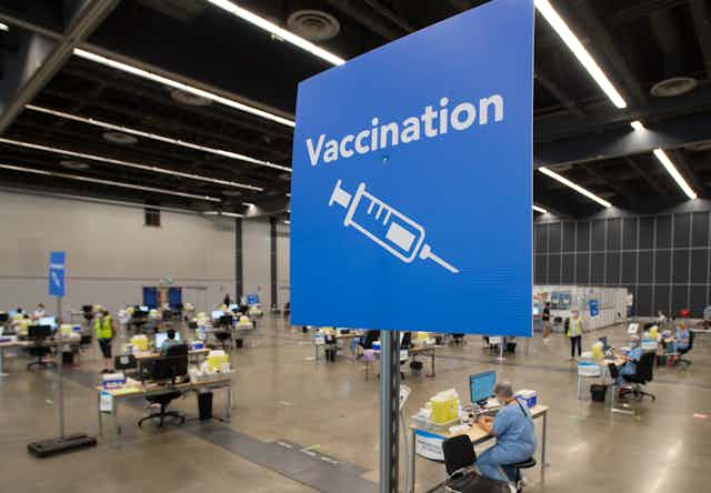 A blue sign in the foreground reading "Vaccination" with an illustration of a syringe, in front of a large space where health-care workers are setting up vaccination stations