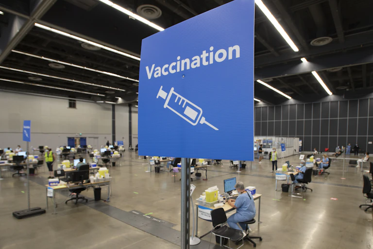 A blue sign in the foreground reading "Vaccination" with an illustration of a syringe, in front of a large space where health-care workers are setting up vaccination stations
