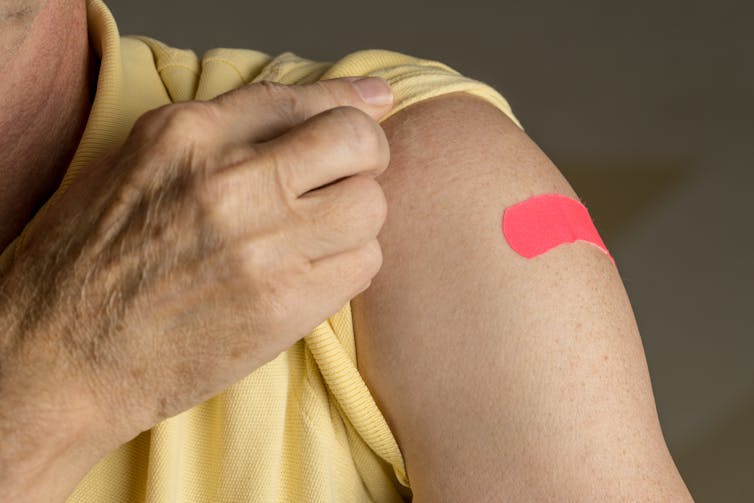 An older person in a yellow top pulls their sleeve up to reveal a pink bandaid.