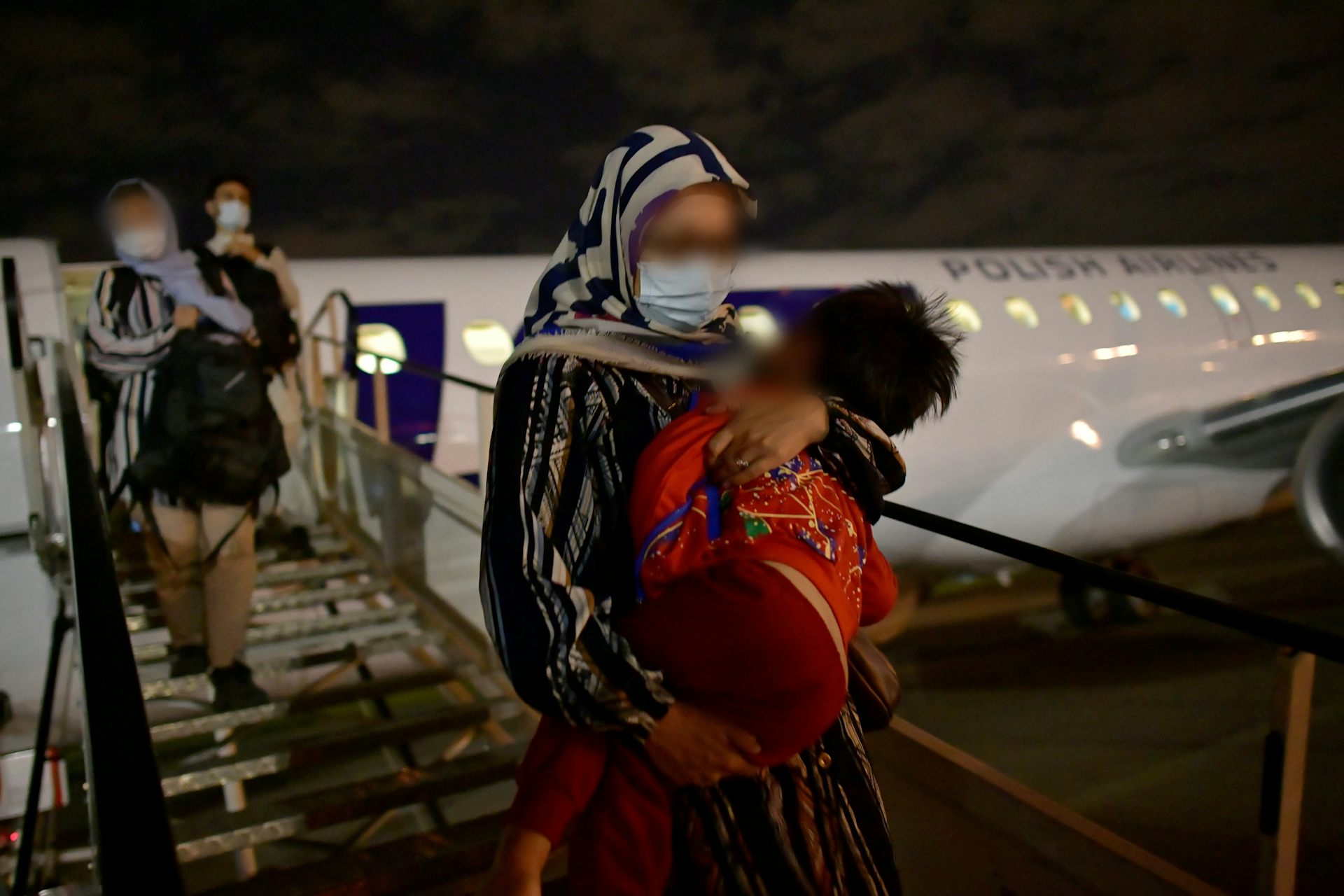 Women with their faces pixelated out walk down the ramp from an aircraft, one carrying a small child.