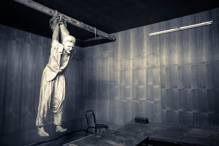 A figure of a person suspended by the arms from a bar across a room, with their feet unable to reach the ground.