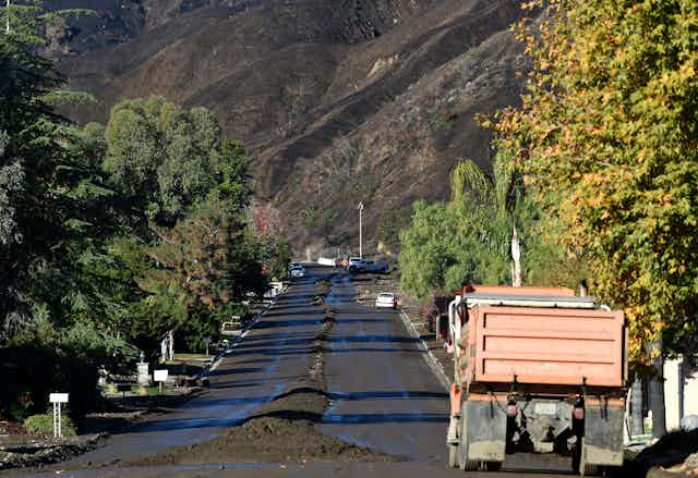 A construction truck moves down a street where workers have pushed debris from a mudflow into the center. A burn-scarred mountainside is in the background