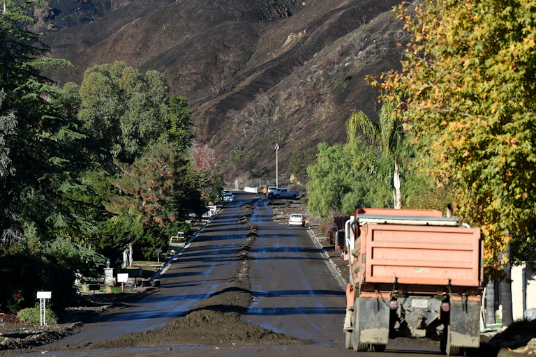 A construction truck moves down a street where workers have pushed debris from a mudflow into the center. A burn-scarred mountainside is in the background