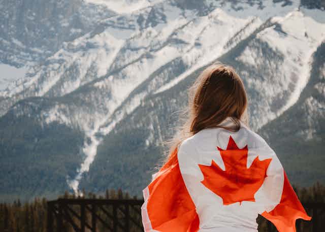 Woman stands facing snow covered mountain ranges with a Canada flag draped over her back
