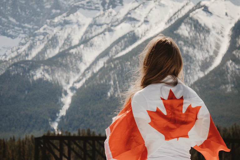 Woman stands facing snow covered mountain ranges with a Canada flag draped over her back