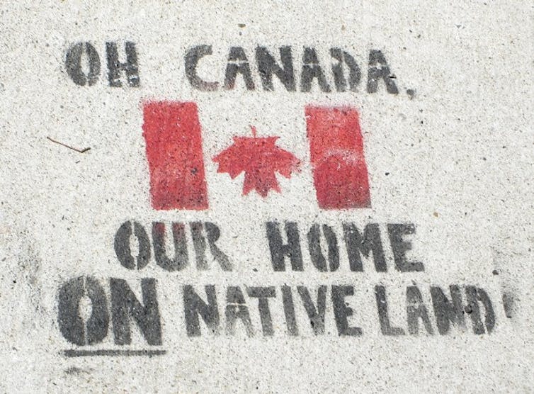 A sign is spray painted on the ground it reads 'Oh Canada, our home ON native land' an upside down Canada flag is also displayed