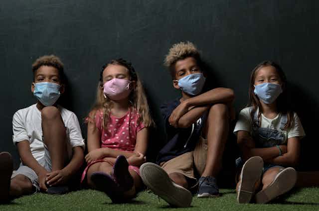 a row of four children wearing surgical masks and sitting on the floor.