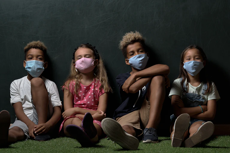 a row of four children wearing surgical masks and sitting on the floor.