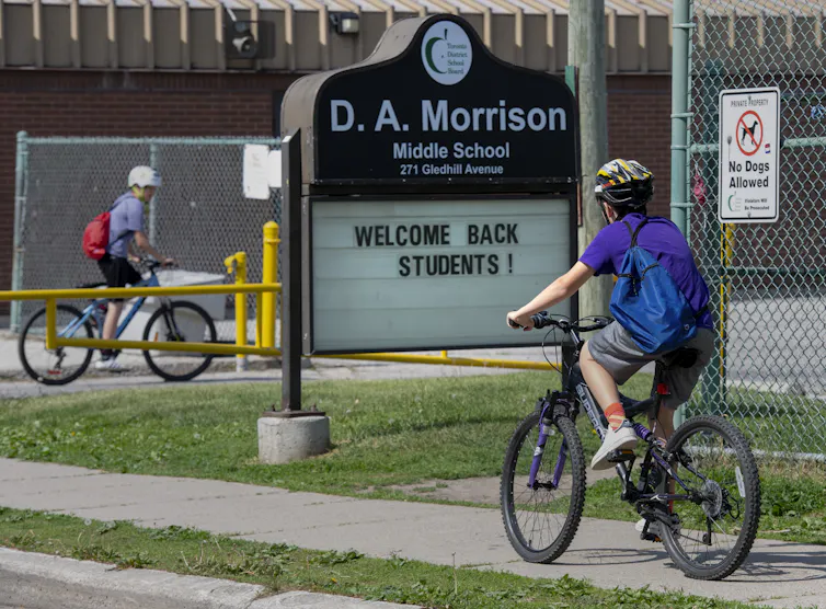 Children riding their bikes into a school with the sign WELCOME BACK STUDENTS