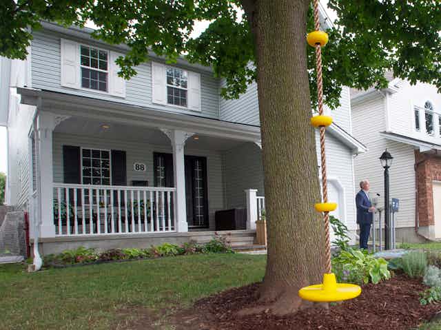 A tree with a yellow swing is positioned infront of a light blue house which has a big white front porch. Erin O'Toole stands on the far right corner.